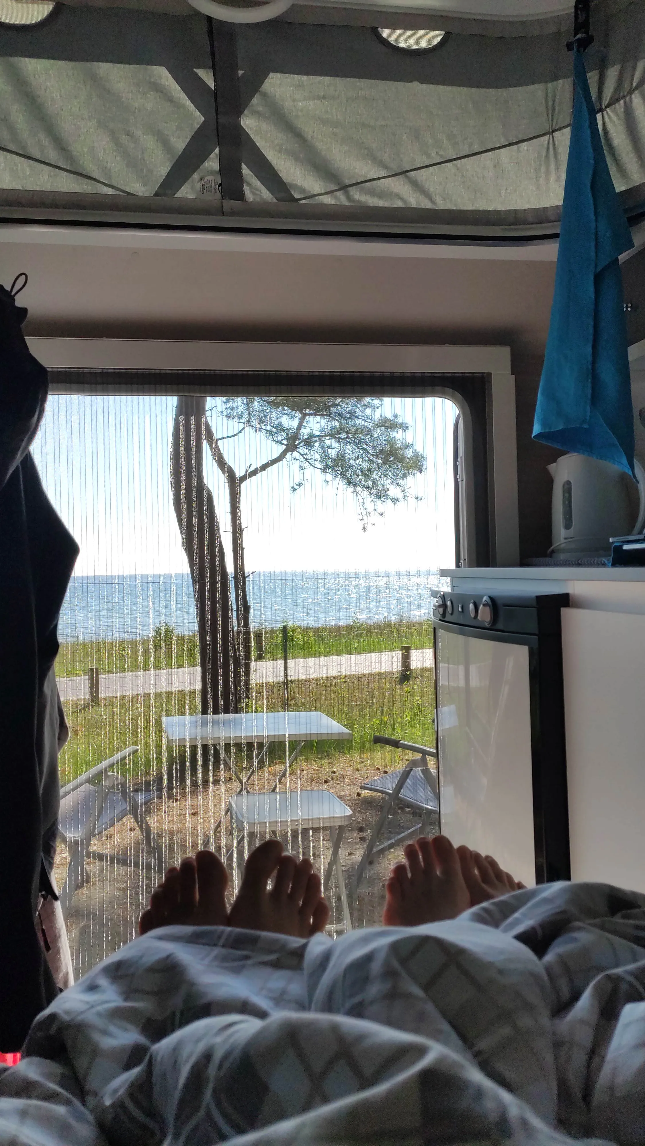 Beim Camping im Caravan, Füße ragen unter der Bettdecke hervor, Wohnwagentür offen mit Blick auf das Meer